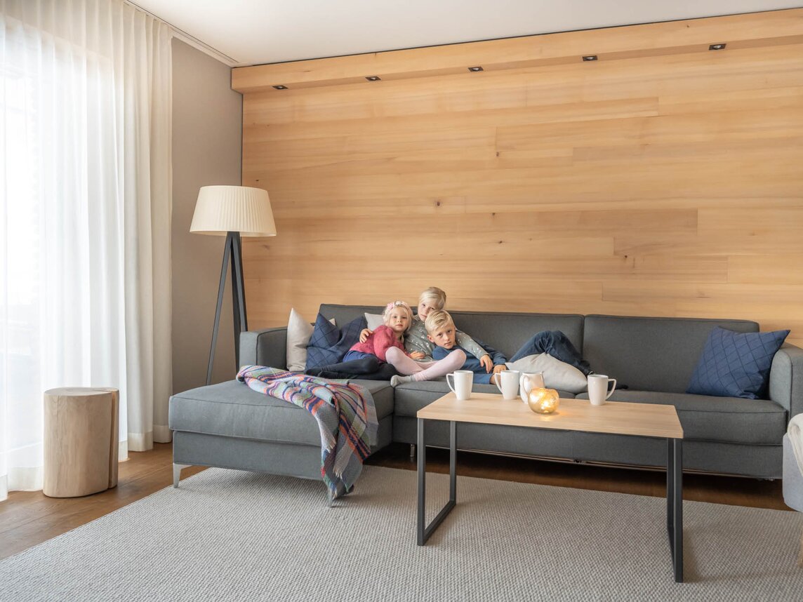 Drei Kinder kuscheln gemeinsam auf einer grauen großen Couch im Wohnzimmer vom Chalet | © Silvretta Montafon - blateral
