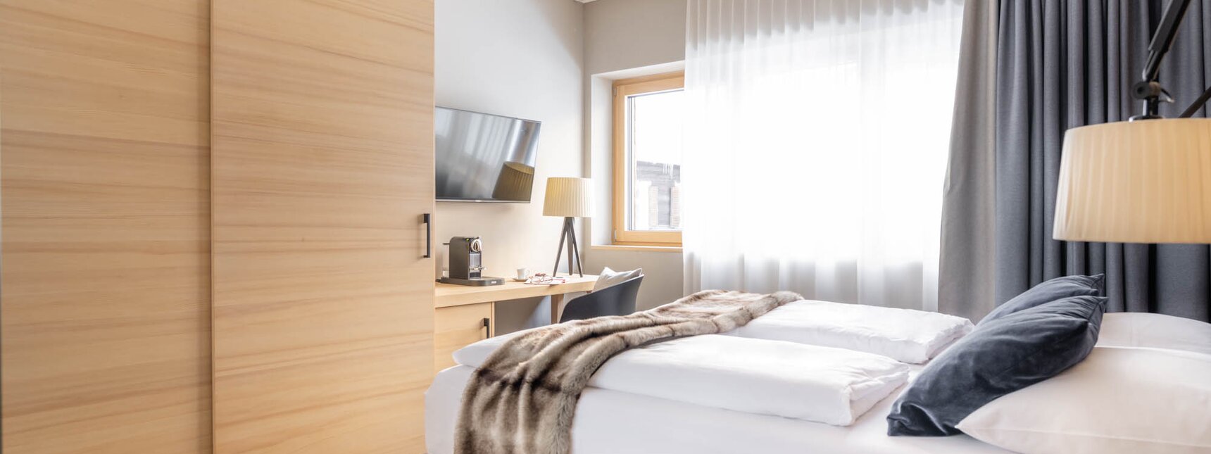 Ein Doppelbett mit grauer Polsterung und weißem Bettbezug und einer Felldecke drüber | © Silvretta Montafon - blateral