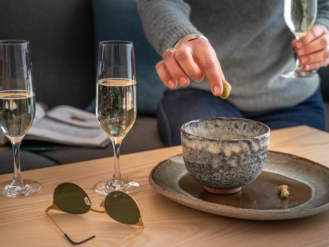 Zwei gefüllte Proseccogläser, eine Sonnenbrille und eine Schale mit Oliven stehen auf dem Tisch und eine Frau sitzt im Hintergrund mit einem Glas und einer Olive in der Hand | ©  Silvretta Montafon - blateral