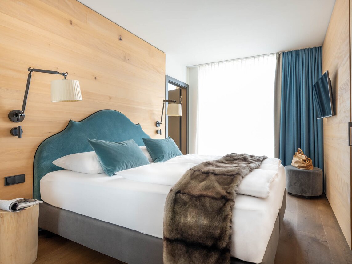 Ein großes Bett mit einer samtigen türkisen Polsterung und einem Fell auf der weißen Bettdecke | © Silvretta Montafon - blateral