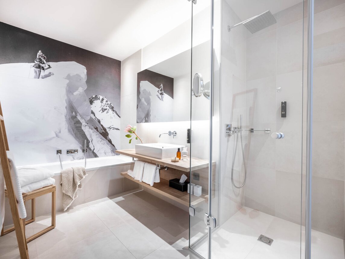 Ein Badezimmer mit einem Waschtisch, einer Dusche und einer Badewanne mit einem Skibild dahinter an der Wand | © Silvretta Montafon - blateral