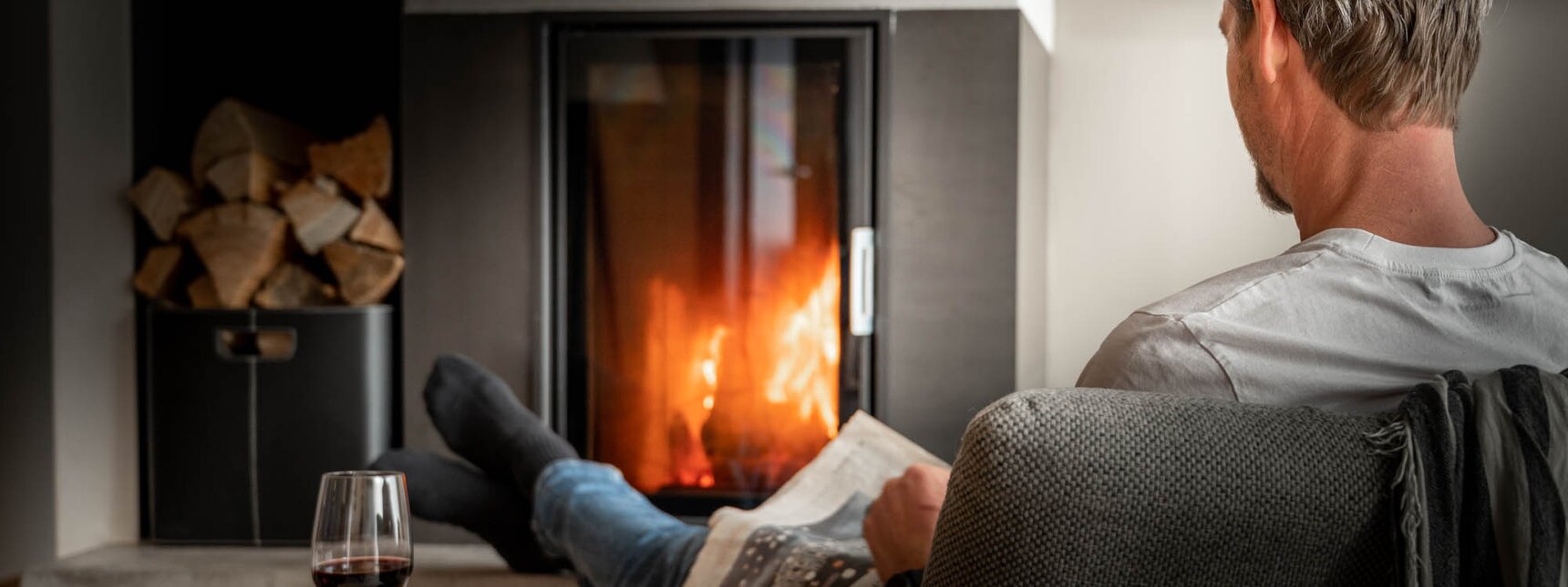 Ein Mann sitzt vor dem Feuer im Kamin, liest seine Zeitung und trinkt Rotwein | © Silvretta Montafon - blateral
