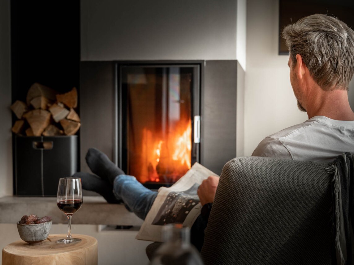 Ein Mann sitzt vor dem Feuer im Kamin, liest seine Zeitung und trinkt Rotwein | © Silvretta Montafon - blateral