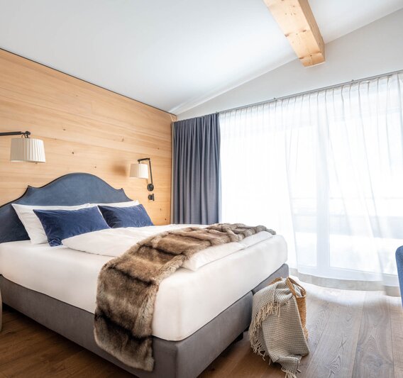 Ein gemütliches großes Bett mit grauer Polsterung und einer Felldecke darüber | © Silvretta Montafon - blateral