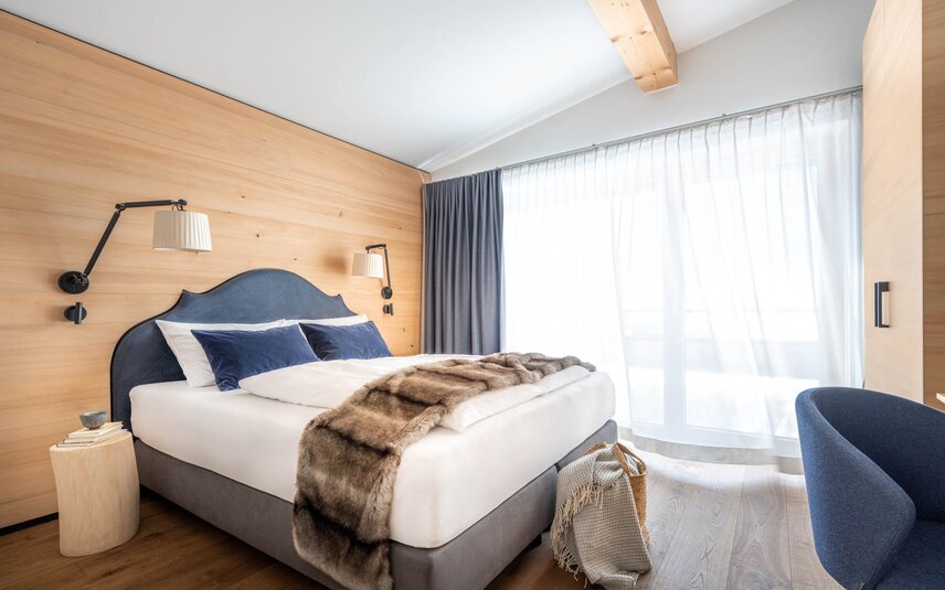 Chalet Sporthotel Silvretta Montafon | © Silvretta Montafon - blateral Ein gemütliches großes Bett mit grauer Polsterung und einer Felldecke darüber | © Silvretta Montafon - blateral
