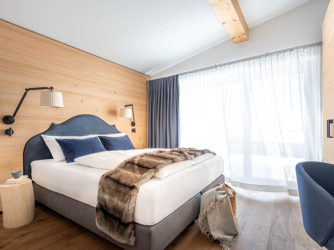 Ein gemütliches großes Bett mit grauer Polsterung und einer Felldecke darüber | © Silvretta Montafon - blateral