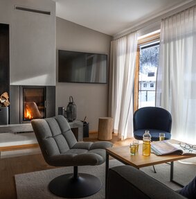 Ein gemütlicher Wohnbereich mit einem brennenden Feuer im Kamin, zwei gepolsterten Stühlen und Getränken auf dem Tischlein | © Silvretta Montafon - blateral