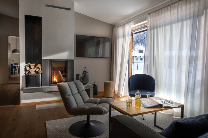 Chalet Sporthotel Silvretta Montafon | © Silvretta Montafon - blateral Ein gemütlicher Wohnbereich mit einem brennenden Feuer im Kamin, zwei gepolsterten Stühlen und Getränken auf dem Tischlein | © Silvretta Montafon - blateral
