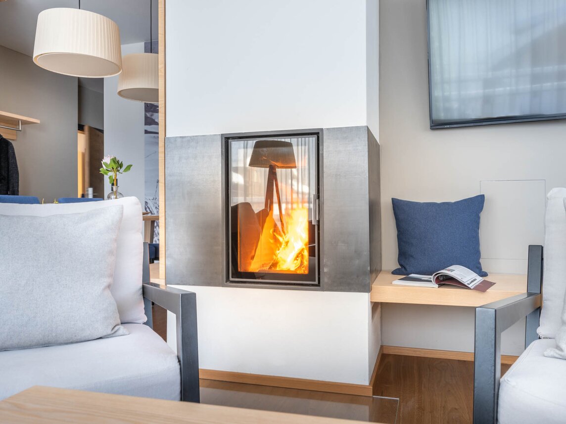 Ein brennendes Feuer im Kamin, ein blauer Polster auf der Kaminbank und eine graue Couch | © Silvretta Montafon - blateral