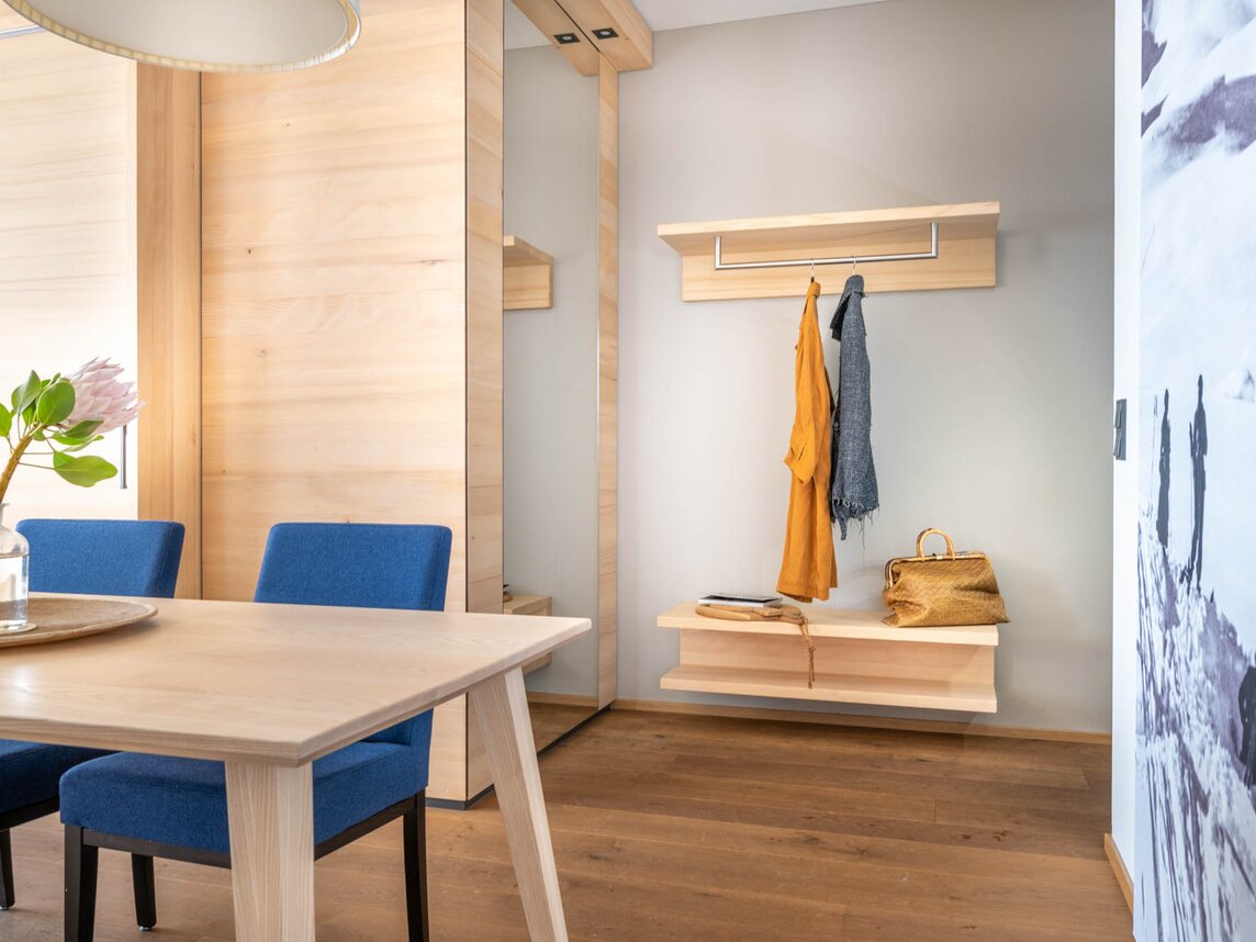 Eine Garderobe aus Holz mit zwei Jacken, ein schwarz-weiß Bild an der Wand und einem Esstisch mit blauen Stühlen | © Silvretta Montafon - blateral
