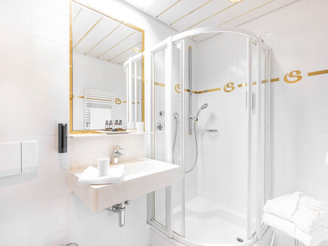 Ein Badezimmer mit weißen Fließen und einem Spiegel in Goldoptik und einer Dusche | © Silvretta Montafon - blateral